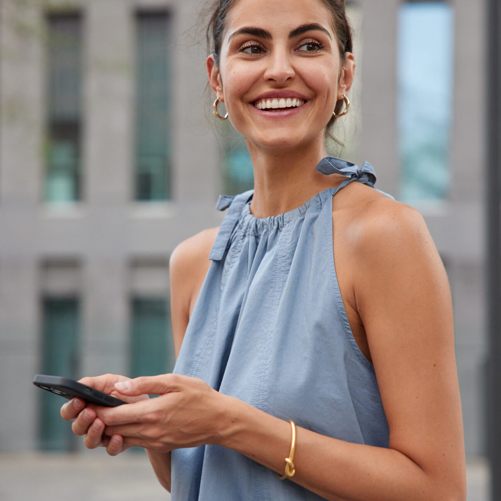 Vertical shot of pleased happy woman smiles toothily holds mobile phone in hands creats travel publication and shares to social media work glad to read received messahe wears blue t tshirt sunglasses